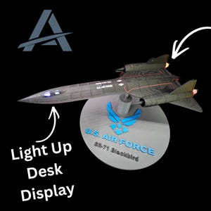 Puede incluir: Un modelo de avión SR-71 Blackbird negro montado en una base circular con el logotipo y texto de la U.S. Air Force. El modelo incluye ventanas de cabina y escapes de motor iluminados, con el texto Light Up Desk Display visible.