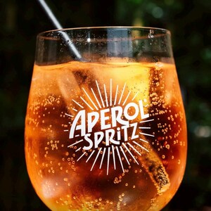 May include: A glass of Aperol Spritz cocktail with ice cubes and a black straw. The drink is a vibrant orange color, with the words "Aperol Spritz" in white, stylized text. The background is blurred, with green foliage.