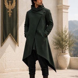 Può includere: Un cappotto verde scuro a maniche lunghe con orlo asimmetrico, collo alto e fibbia laterale. Il cappotto è indossato sopra pantaloni scuri e stivali di pelle. Un logo circolare nell'angolo riporta la scritta PHANTOMEFORGE CRAFTS.