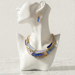 Peut inclure: Un collier géométrique bleu, doré et blanc et une paire de boucles d'oreilles assorties présentés sur un buste de mannequin blanc. Le collier présente une série de formes rectangulaires et angulaires.
