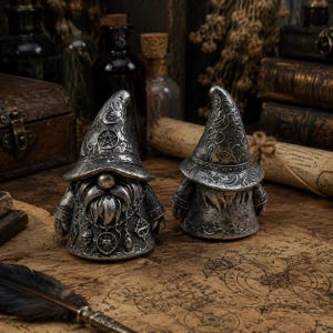 May include: Two silver-toned figurines of bearded figures wearing pointed hats. The left figure features a crescent moon and pentagram on the hat, with a chain necklace holding small charms. The right figure displays intricate swirling patterns.