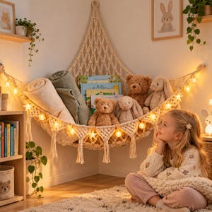 May include: A cream-colored macrame corner hammock suspended on a wall, filled with two rolled blankets, three children's books, two teddy bears, and two plush rabbits. The hammock is decorated with a string of glowing star-shaped fairy lights.