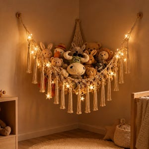 May include: A cream-colored macrame corner hammock for stuffed animals, featuring a knotted net design with hanging tassels along the bottom edge. The hammock is illuminated by a string of warm white star-shaped lights woven into the cord.