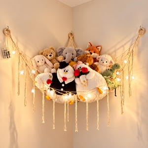 May include: A beige macrame corner hammock for stuffed animals, suspended by three wooden rings. The net holds various plush toys, including a bear, elephant, fox, and snowman, and is decorated with a string of glowing star-shaped lights.
