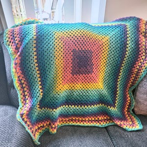 Op de afbeelding: Een vierkante gehaakte deken met een veelkleurig granny square patroon. Het ontwerp verloopt in concentrische lagen van donkerpaars, felgeel, koraalroze, talingblauw en lichtgroen, wat zorgt voor een levendig kleurverloop over het geheel.