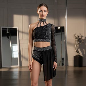 Puede incluir: Traje de baile negro con adornos de pedrería. El conjunto presenta un top corto de un solo hombro, una manga larga y un pantalón corto de cintura alta con una superposición de falda. El choker y las cinturas también están adornados con pedrería.