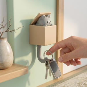 Könnte beinhalten: Ein beiger Wand-Schlüsselhalter mit einer grauen Katzenfigur, die aus einer Box hervorschaut. Der Schlüsselhalter hat einen gebogenen Haken zum Aufhängen von Schlüsseln. Die Katze hat eine kleine schwarze Nase und Augen.