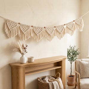 Op de afbeelding: Een crèmekleurige macramé-banner met driehoekige vormen en franjes hangt boven een houten console. De banner is versierd met houten kralen. Een witte vaas met droogbloemen, een kaars en een potplant staan op de tafel.