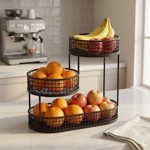 Könnte beinhalten: Ein dreistufiger Obstkorb aus dunklem Metall, gefüllt mit Orangen, Äpfeln und Bananen. Der mehrstufige Korb steht auf einer weißen Arbeitsplatte. Im Hintergrund befindet sich eine Kaffeemaschine. Rechts liegt eine gefaltete Serviette.
