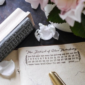 May include: A vintage-style journal with a brown leather cover and gold accents. The journal is open to a page with a calendar printed on it. The calendar is labeled "The Journal of Alice Thornburg" and includes the months of the year, days of the week, and numbers 1 through 31. A gold pen is lying on the open journal.