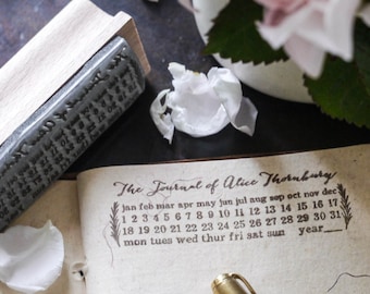 May include: A vintage-style journal with a brown leather cover and gold accents. The journal is open to a page with a calendar printed on it. The calendar is labeled "The Journal of Alice Thornburg" and includes the months of the year, days of the week, and numbers 1 through 31. A gold pen is lying on the open journal.