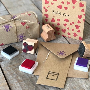 以下が含まれることがあります： ハート、手形、そして「With Love」と「Handmade by」の文字を含む様々なデザインのスタンプセット。スタンプは、様々な色のインクパッドと共に、茶色の紙の表面に配置されています。
