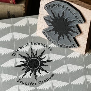 May include: A wooden stamp with a sunburst design and the words "This book belongs to Jennifer Graham" is next to a matching sunburst design on a gray book. The stamp is on a wooden block.