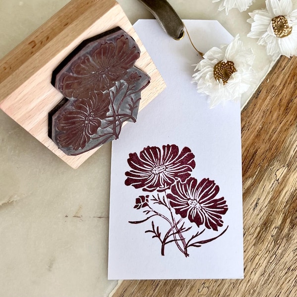 May include: A wooden stamp with a floral design is shown next to a stamped image of the same flowers on a white tag. The stamp and the stamped image are a deep burgundy color. Dried white flowers with gold centers are scattered around the tag.