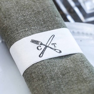 Peut inclure: Une serviette en lin verte avec une bande blanche sur laquelle est dessinée en noir et blanc une fourchette et un couteau avec les lettres "C" et "T".