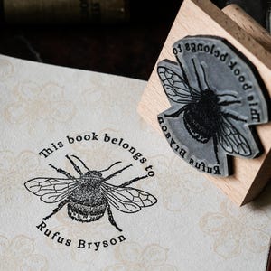 May include: A wooden stamp featuring a bee design and the text "This book belongs to Rufus Bryson" is pressed onto floral paper. Another stamp with a bee design is also visible.
