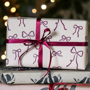 May include: Three wrapped gifts stacked. The top gift is wrapped in light paper with a repeating bow pattern and tied with a burgundy ribbon. The middle gift is wrapped in brown paper with a bird and musical note design.
