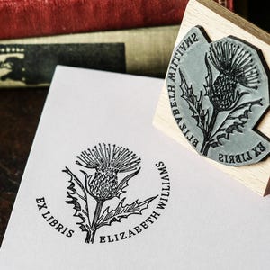 May include: A wooden stamp with a thistle design and the text "EX LIBRIS ELIZABETH WILLIAMS" is shown next to a stamped image of the same design on white paper. The stamp is gray and black, and the paper is white. The background includes old books.