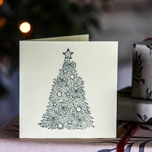 Op de afbeelding: Een vierkante kerstkaart met een groene illustratie van een kerstboom versierd met een ster, strikken en ballen. De lichtbeige kaart ligt op een bruin oppervlak, met verpakte cadeaus op de achtergrond. De kaart is ongeveer 14 cm x 14 cm groot.