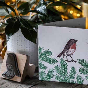 May include: A greeting card featuring a printed illustration of a robin with a red breast, perched on a green fir branch design. A wooden stamp with the robin image is also visible. The card is on a wooden surface.