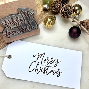 以下が含まれることがあります： バーガンディ色の筆記体で「Merry Christmas」と書かれた白いタグ。同じテキストの木製スタンプが見えます。画像にはクリスマスオーナメントと松ぼっくりも含まれており、ホリデーテーマを示唆しています。