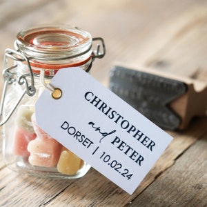 Op de afbeelding: Een klein glazen potje gevuld met kleurrijke snoepjes, vastgebonden met een wit label waarop staat "Christopher and Peter Dorset | 10.02.24".