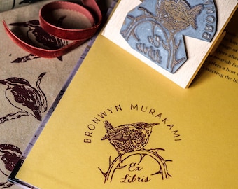 Può includere: Un libro giallo con un'illustrazione di un uccello marrone e bianco sulla copertina. Il libro ha un timbro di gomma marrone con il testo "Bronwyn Murakami Ex Libris" e una piccola illustrazione di un uccello. Il timbro è su un blocco di legno con il testo "Maiarum Hymno BBO" in un design circolare.