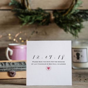 May include: A white card with black calligraphy that reads "12.07.18" and "PLEASE SAVE THE DATE FOR THE WEDDING OF LUCY CHANDLER & BEN HULL IN LONDON". A red heart with a sunburst design is below the text.