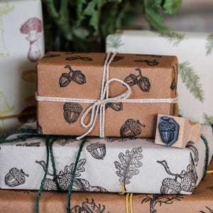 May include: Several gift-wrapped parcels with acorn and oak leaf designs. The packages are wrapped in brown and white paper, tied with twine, and feature black ink illustrations. A wooden stamp with an acorn design is also visible.