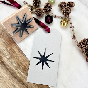 Könnte beinhalten: Ein Holzstempel mit einem Sternmuster neben einem Geschenkanhänger mit demselben Sternmotiv. Der Stempel hat einen dunkelgrauen Stern, der an einem hellbraunen Holzblock befestigt ist. Der Geschenkanhänger ist hellgrau mit einem schwarzen Stern. Dekorative Tannenzapfen und Ornamente befinden sich im Hintergrund.