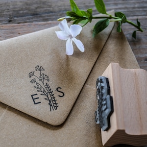 以下が含まれることがあります： 花柄のデザインが施された茶色の紙の封筒に「E.S.」のイニシャルが押印されています。封筒の隣には、花柄の刻印が施された木製のスタンプがあります。