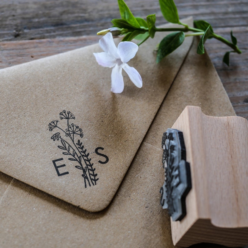 以下が含まれることがあります： 花柄のデザインが施された茶色の紙の封筒に「E.S.」のイニシャルが押印されています。封筒の隣には、花柄の刻印が施された木製のスタンプがあります。