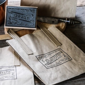 K&ouml;nnte beinhalten: Drei braune Papiert&uuml;ten mit schwarzen Stempeln. Die Stempel zeigen den Text "Farro", "Borlotti Beans" und "Ellini Beans". Jeder Stempel enth&auml;lt den Text "Fattorie Del Tratturo" und dekorative Verzierungen.