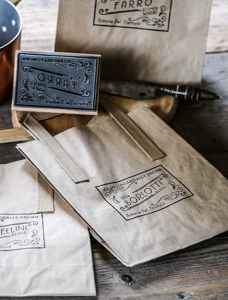 K&ouml;nnte beinhalten: Drei braune Papiert&uuml;ten mit schwarzen Stempeln. Die Stempel zeigen den Text "Farro", "Borlotti Beans" und "Ellini Beans". Jeder Stempel enth&auml;lt den Text "Fattorie Del Tratturo" und dekorative Verzierungen.
