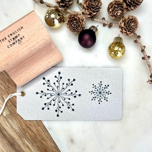Puede incluir: Un sello de madera de The English Stamp Company junto a una etiqueta con dos diseños de copos de nieve. La etiqueta es de color gris claro con diseños de copos de nieve azul oscuro. Adornos navideños y piñas en el fondo.