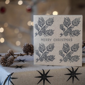 Peut inclure: Carte de Noël carrée en papier kraft avec quatre illustrations de branches de pin et de pommes de pin. Le texte « MERRY CHRISTMAS » est imprimé en noir. La carte est posée sur du papier d'emballage à motifs d'étoiles.