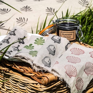 May include: A wicker picnic basket with a white tablecloth and three linen napkins with different printed designs. The napkins feature images of leaves, acorns, and seashells. A jar of elderberry chutney is on top of the basket.
