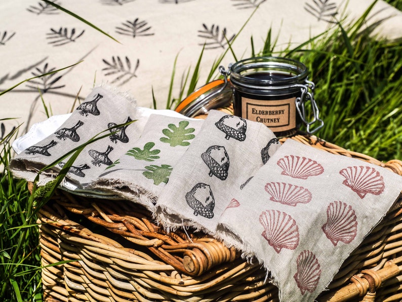 May include: A wicker picnic basket with a white tablecloth and three linen napkins with different printed designs. The napkins feature images of leaves, acorns, and seashells. A jar of elderberry chutney is on top of the basket.