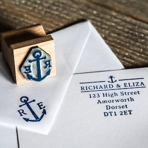 Puede incluir: Un sello de madera con un diseño de ancla azul y las letras "R" y "E" se muestra junto a un sobre sellado. El sobre tiene el nombre "RICHARD & ELIZA" y la información de la dirección impresa en azul.