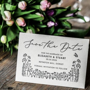 以下が含まれることがあります： 「Save the Date」のカードで、花柄の縁取りと「Save the Date」の文字が筆記体で書かれています。カードには、「For the marriage of Elizabeth & Stuart 20.05.2023 Keynston Mill, Dorset Formal invitation to follow.」という文章も書かれています。