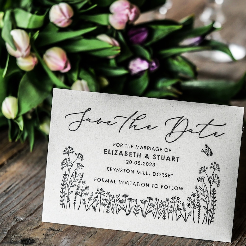 以下が含まれることがあります： 「Save the Date」のカードで、花柄の縁取りと「Save the Date」の文字が筆記体で書かれています。カードには、「For the marriage of Elizabeth & Stuart 20.05.2023 Keynston Mill, Dorset Formal invitation to follow.」という文章も書かれています。
