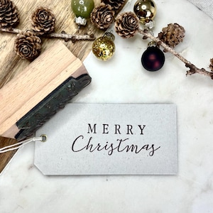 以下が含まれることがあります： 「MERRY Christmas」と書かれたタグが特徴的な、お祝いのイメージ。木製のスタンプ、松ぼっくり、クリスマスオーナメントが写っています。オーナメントは金色、緑色、ブルゴーニュ色です。タグは白い紐で取り付けられています。