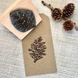 Może przedstawiać: Drewniana pieczątka z motywem szyszki, obok odbitego obrazu tej samej szyszki na brązowej etykiecie. Trzy małe szyszki i gałązka znajdują się w tle. Pieczątka ma jasny drewniany uchwyt.