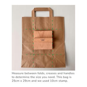 K&ouml;nnte beinhalten: Eine braune Papiert&uuml;te mit einem Muster aus gr&uuml;nen gestrichelten Linien. Die Tasche ist gefaltet, um die Gr&ouml;&szlig;e eines 10 cm gro&szlig;en quadratischen Stempels zu zeigen. Der Stempel ist aus Holz und tr&auml;gt den Text "THE ENGLISH STAMP COMPANY". Messen Sie zwischen den Falten, Knickfalten und Griffen, um die ben&ouml;tigte Gr&ouml;&szlig;e zu ermitteln. Diese Tasche ist 25 cm x 29 cm gro&szlig; und wir haben einen 10 cm gro&szlig;en Stempel verwendet.