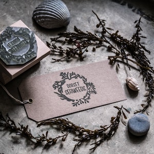 May include: A brown paper tag with the text "Dorset Seaweeds" in a black circle with a floral design. The tag is on a gray surface with a seashell, a wooden stamp, and dried seaweed.