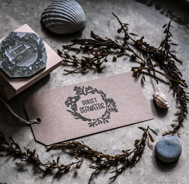 May include: A brown paper tag with the text "Dorset Seaweeds" in a black circle with a floral design. The tag is on a gray surface with a seashell, a wooden stamp, and dried seaweed.