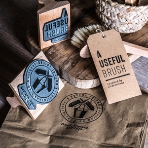 May include: A set of three wooden stamps with black ink. The stamps feature the text "A Useful Brush" and a design of a bucket and brush. The stamps are on a brown paper bag with a brown tag that reads "A Useful Brush Inspired by Generations".