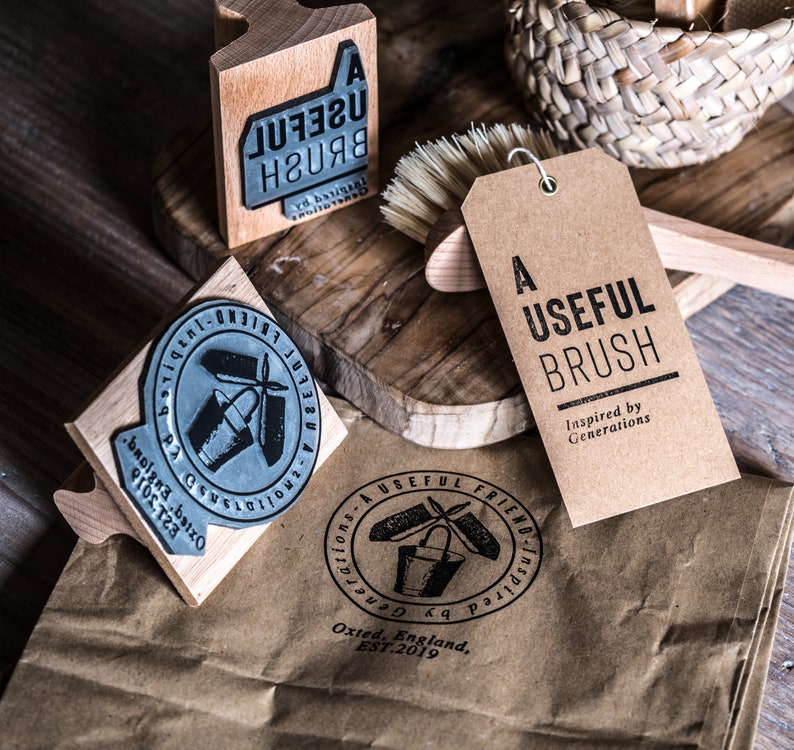 May include: A set of three wooden stamps with black ink. The stamps feature the text "A Useful Brush" and a design of a bucket and brush. The stamps are on a brown paper bag with a brown tag that reads "A Useful Brush Inspired by Generations".