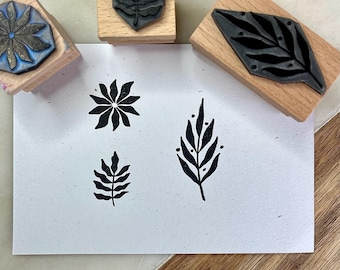 May include: A collection of wooden stamps with botanical designs. The stamps are made of wood and feature various leaf and floral patterns. The stamped images are in black ink on white paper. The stamps are arranged on a light-colored surface.