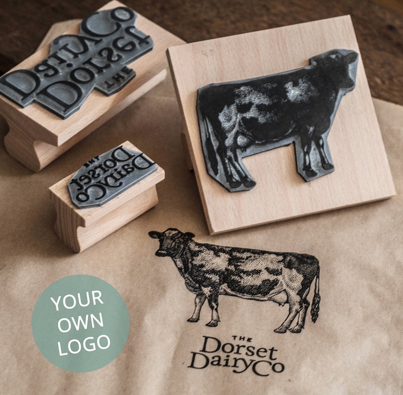 K&ouml;nnte beinhalten: Drei Holzstempel mit verschiedenen Designs. Der erste Stempel zeigt den Text "The Dorset Dairy Co." in einer stilisierten Schriftart. Der zweite Stempel zeigt eine schwarz-wei&szlig;e Silhouette einer Kuh. Der dritte Stempel zeigt den Text "Your Own Logo" in einem Kreis.